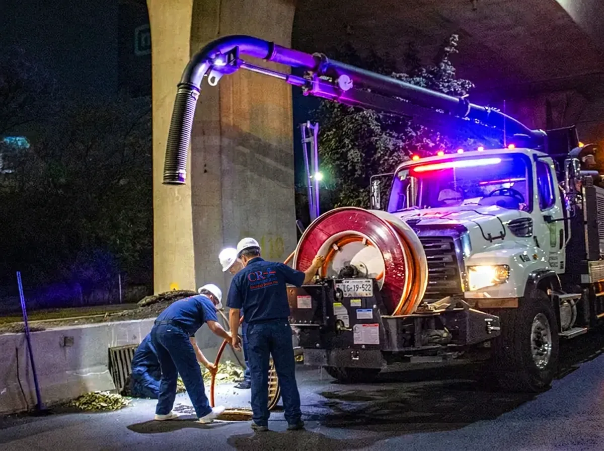Emergency drain crew working under floodlights at night beside a hydro jetting truck with hose reels deployed to a street drain